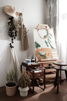 an easel and some plants in front of a painting on the wall next to a window