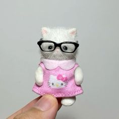 a hand holding a hello kitty doll with glasses on it's head and wearing a pink dress