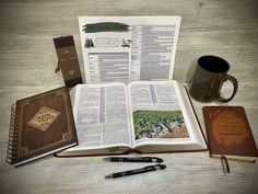 an open book, pen and two coffee mugs on a wooden table with other items