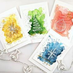 four watercolor christmas cards on a white table with string lights and paper snowflakes