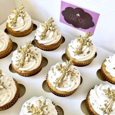 twelve cupcakes with white frosting and gold decorations on top are sitting in a box