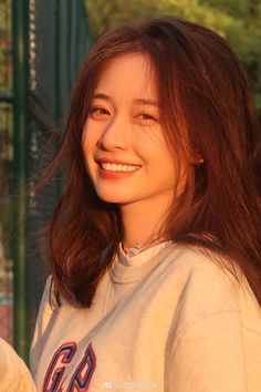 a woman with long brown hair smiling and wearing a white shirt, standing in front of a green fence