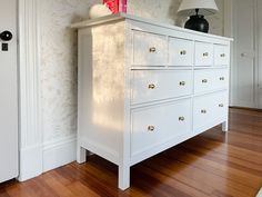 a white dresser sitting on top of a hard wood floor next to a lamp and door