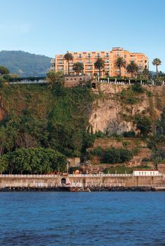 a large building sitting on top of a cliff next to the ocean with trees and bushes in front of it