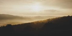 the sun shines brightly through the clouds in the distance as trees stand on top of a hill