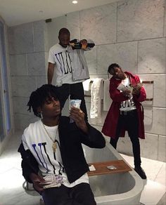 three men in a bathroom taking pictures with their cell phones and one man is drinking