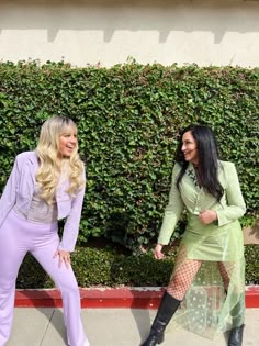 two women dressed in costumes standing next to each other on the sidewalk near a hedge