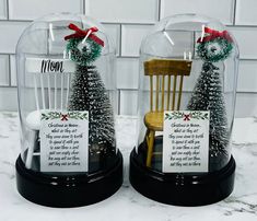 two snow globes sitting on top of a counter with christmas decorations in them and cards attached to the chairs