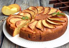 a cake on a white plate with slices cut out and apples in the back ground