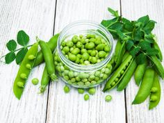 peas and pea pods in a glass bowl on a white wooden table with green leaves