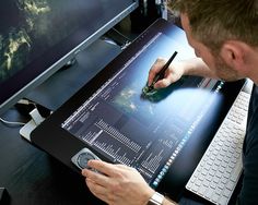 a man writing on a piece of paper next to a computer monitor and keyboard with another person holding a pen