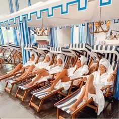 several women in white robes sitting on beach chairs under an umbrella with blue and white stripes