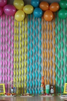 colorful balloons and streamers decorate the wall behind a table with snacks on it in front of an orange, yellow, pink, green, blue, and white backdrop