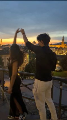 a man and woman standing next to each other on top of a balcony at sunset