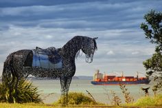 a sculpture of a horse stands in front of the ocean with a cargo ship in the background