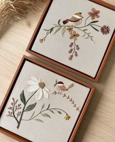 two framed pictures with flowers and birds in them on a wooden table next to a plant