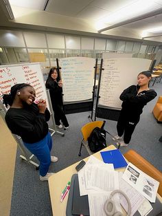 two women standing in front of a whiteboard with writing on it and another woman sitting at a desk