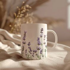 a coffee cup with lavender flowers painted on it sitting on a white tablecloth next to a plant