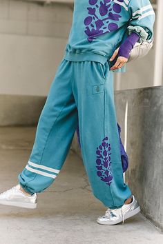 a young boy wearing blue sweatpants and white sneakers, leaning against a wall with his hand on his hip