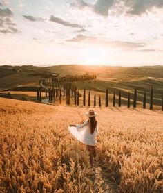 a woman in a white dress and hat walking through a wheat field with the sun setting behind her