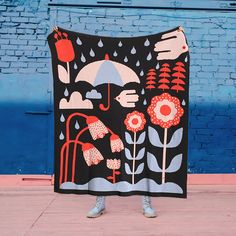 a woman standing on the sidewalk holding up a black blanket with flowers and an umbrella