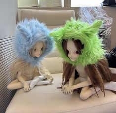 two dolls are sitting on a chair wearing furry hats and wigs, one is green and the other is blue