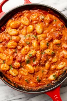 a red skillet filled with pasta and sauce on top of a white marble counter