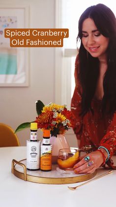 a woman sitting at a table with some bottles and flowers on it, holding an orange