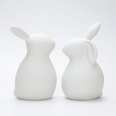 two white ceramic rabbits sitting next to each other on a white surface with one facing the camera