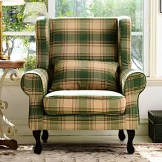 a green plaid chair sitting in front of a window next to a table with a lamp on it