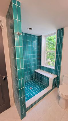 a blue tiled bathroom with a toilet and window in the corner, next to a walk - in shower