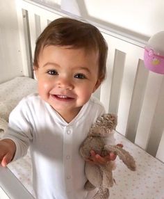 a smiling baby holding a stuffed animal in his hand while standing next to a crib