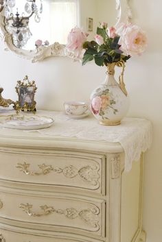 a white dresser with pink flowers in a vase on top and a mirror above it