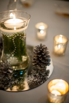 a glass vase filled with pine cones on top of a table next to lit candles