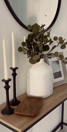 a white vase sitting on top of a wooden table next to two candles and a mirror