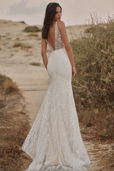 a woman in a white wedding dress standing on a path with bushes and sand behind her