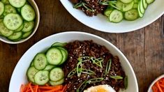 three bowls filled with different types of food on top of a wooden table next to sliced cucumbers and carrots