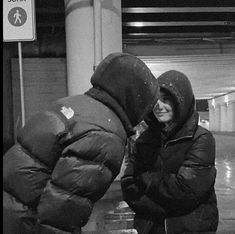 a woman in a hooded jacket standing next to a parking meter and looking at something on the ground