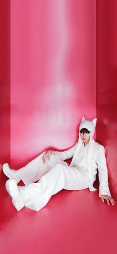 a man in a white suit and hat sitting on a pink floor with his legs crossed