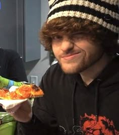 a man holding a slice of pizza while wearing a beanie on top of his head