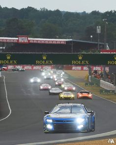 a group of cars driving down a race track with the lights on and people watching