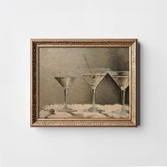 two martini glasses on a table in front of a white wall with a gold frame