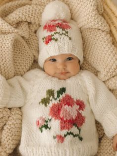 a baby wearing a white knitted sweater and hat with flowers on it in a wicker basket