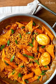 a pan filled with noodles and vegetables on top of a wooden table next to a napkin