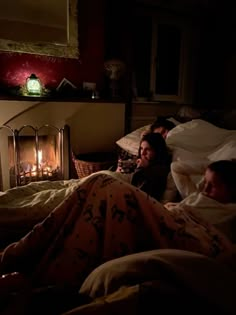 two people are laying in bed next to a fire place with blankets on them and one person is looking at the camera