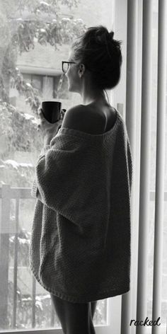 a woman standing in front of a window holding a coffee cup and looking out the window