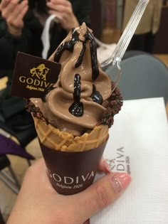 a hand holding a chocolate ice cream sundae in front of a camera and some people