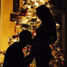 two people standing in front of a christmas tree with lights on it and one person holding the other