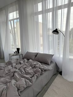 an unmade bed sitting in front of two large windows with sheer curtains on them