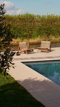 two wooden lounge chairs sitting next to a swimming pool in the middle of a lush green field